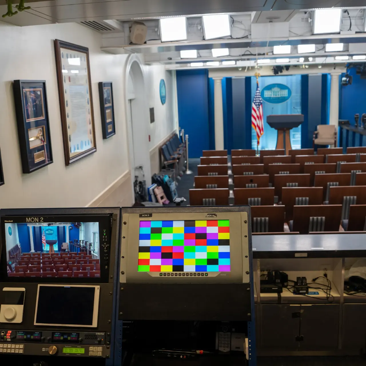 Empty White House briefing room with rows of seats, podium, flag, and monitors.