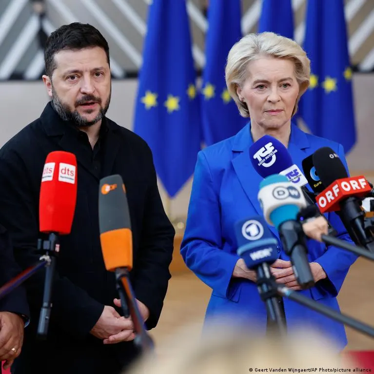 Two officials speak to press microphones before European Union flags, one in a blue blazer.