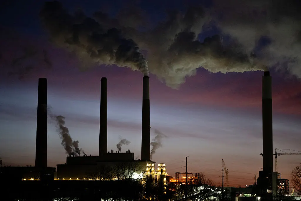 Silhouetted industrial smokestacks emitting plumes of smoke against a dusk sky.