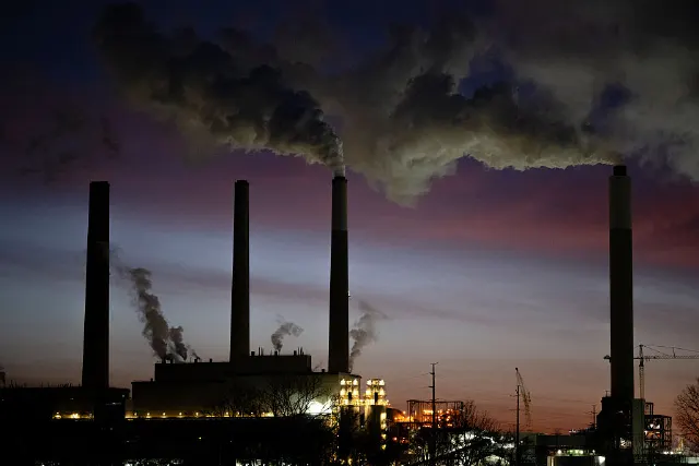 Silhouetted industrial smokestacks emitting plumes of smoke against a dusk sky.