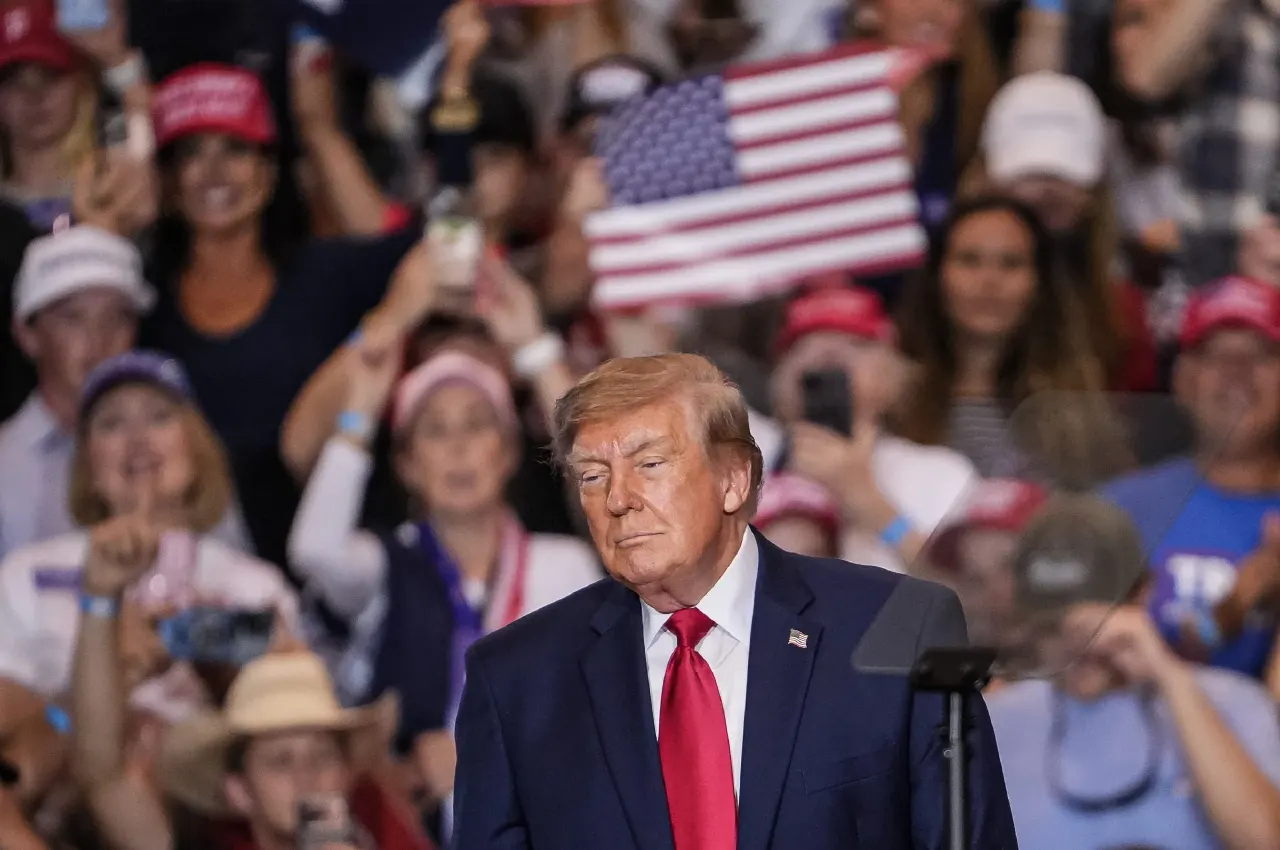Man in navy suit at podium before cheering crowd with red caps and an American flag.
