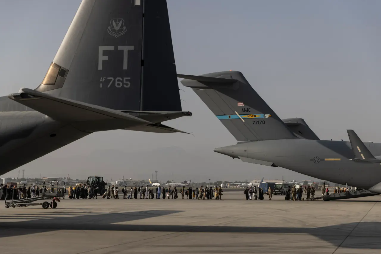 Crowd boarding military transport aircraft on an airfield tarmac, with tails of cargo planes visible.