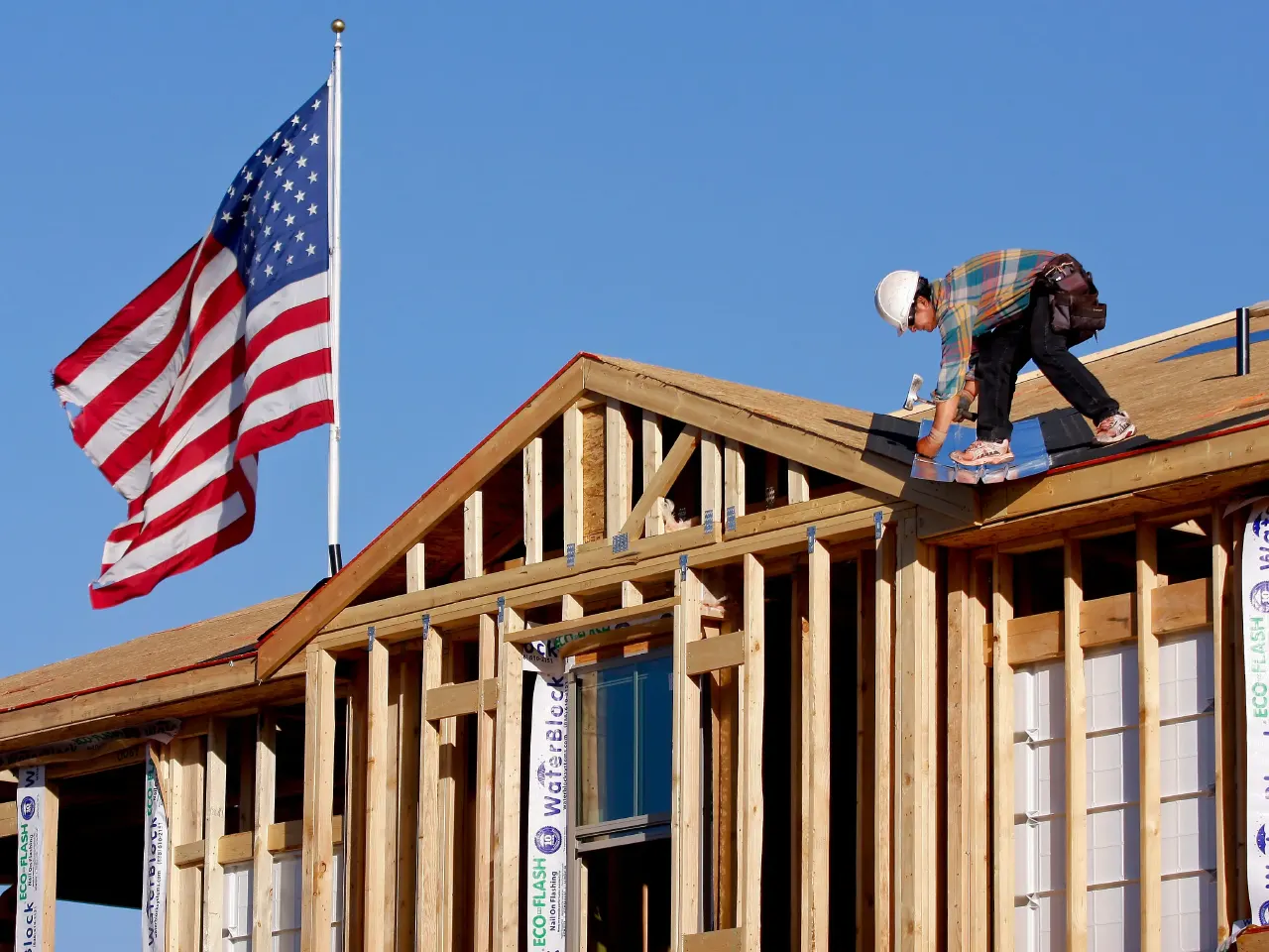 Worker in hard hat and plaid shirt framing a new wood-frame house under construction, with American flag.
