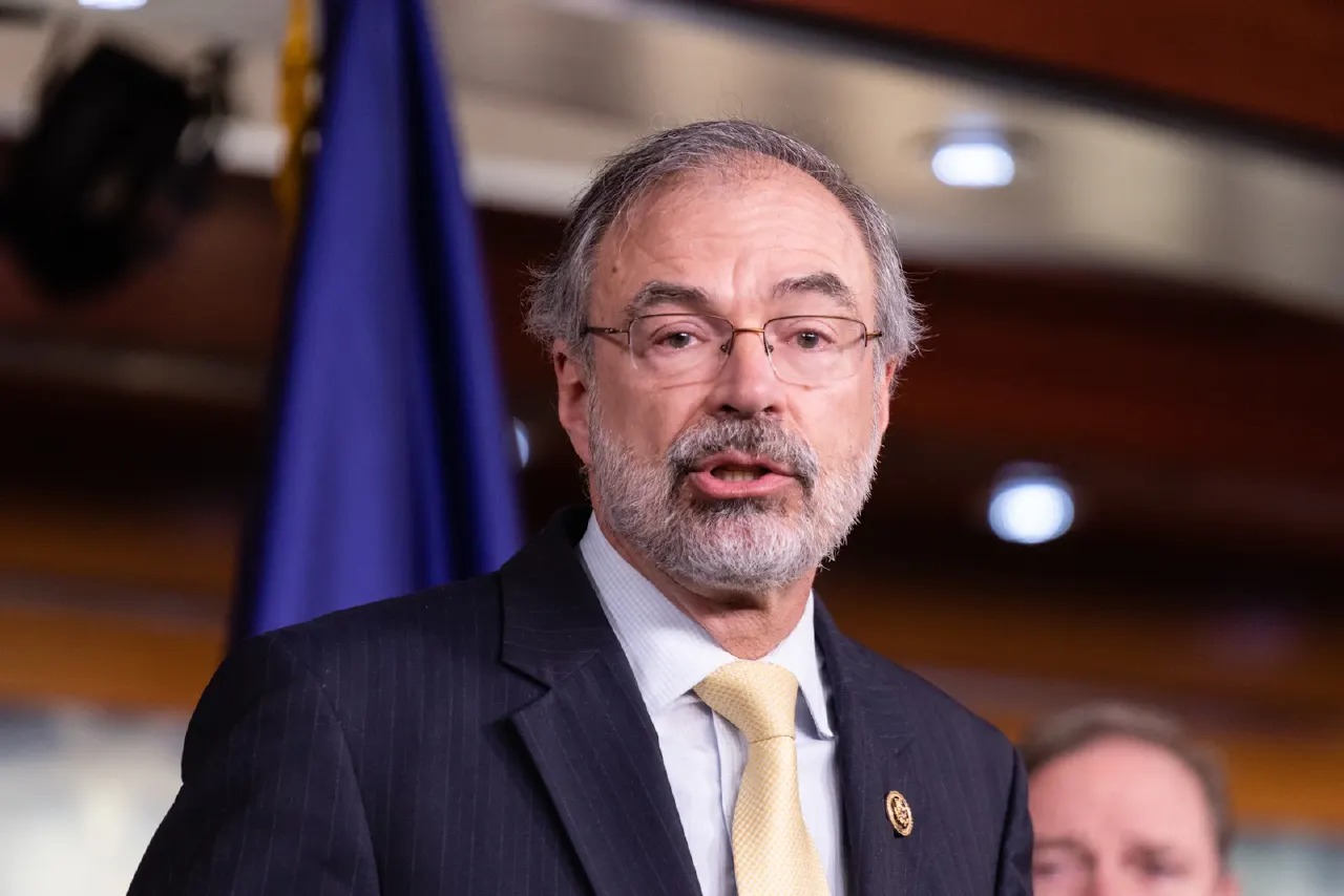 Bearded man with glasses in dark pinstripe suit and yellow tie speaking at press event