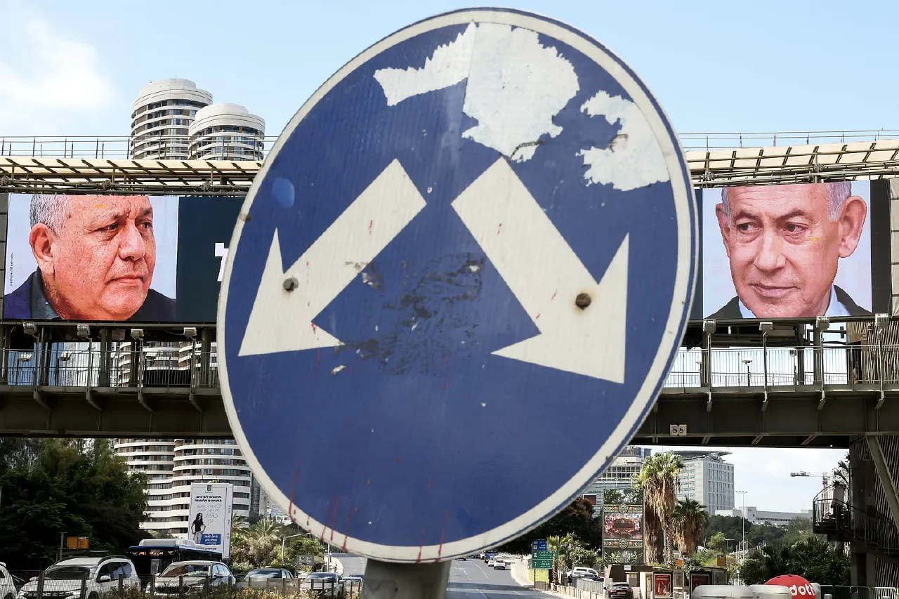Blue road sign in front of large campaign billboards of two men on an overpass, city traffic behind.