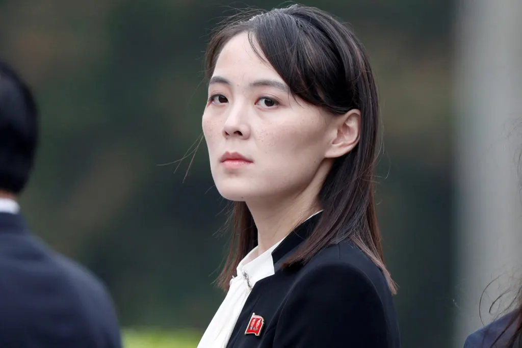Woman in dark jacket with red pin, looking intently sideways outdoors, blurred green background.