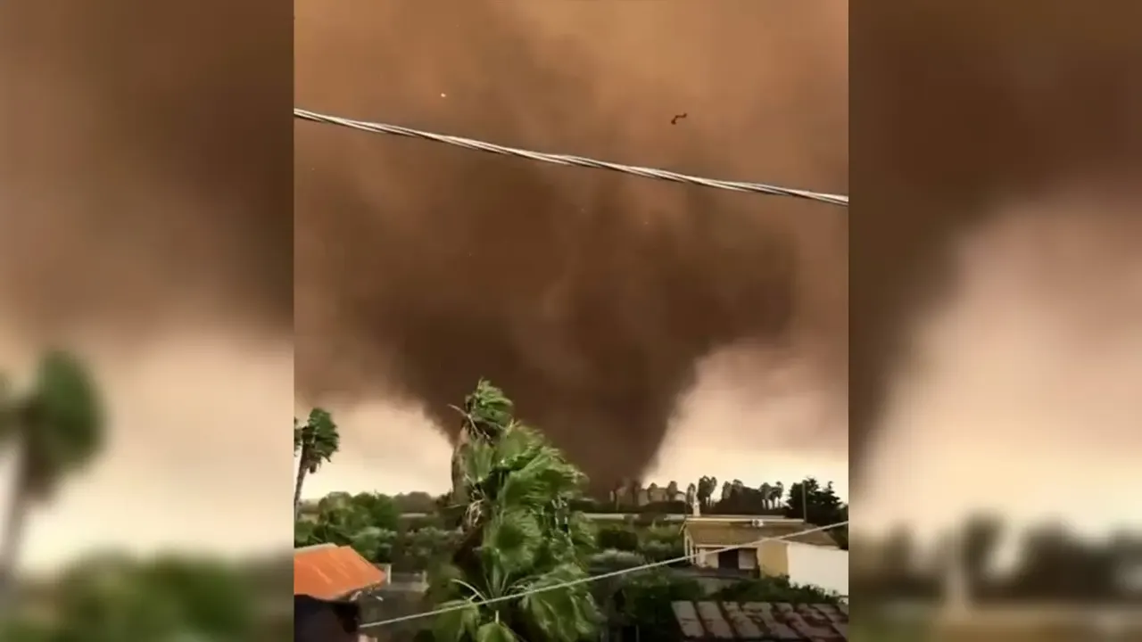 Large dark tornado funnel over houses and palm trees, with power lines in foreground