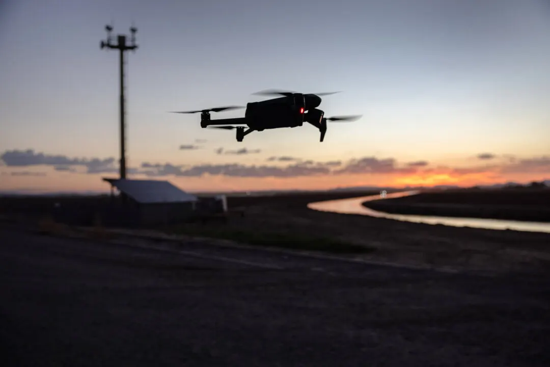 Silhouetted drone hovering over flat desert land at sunset, with a tower and canal.