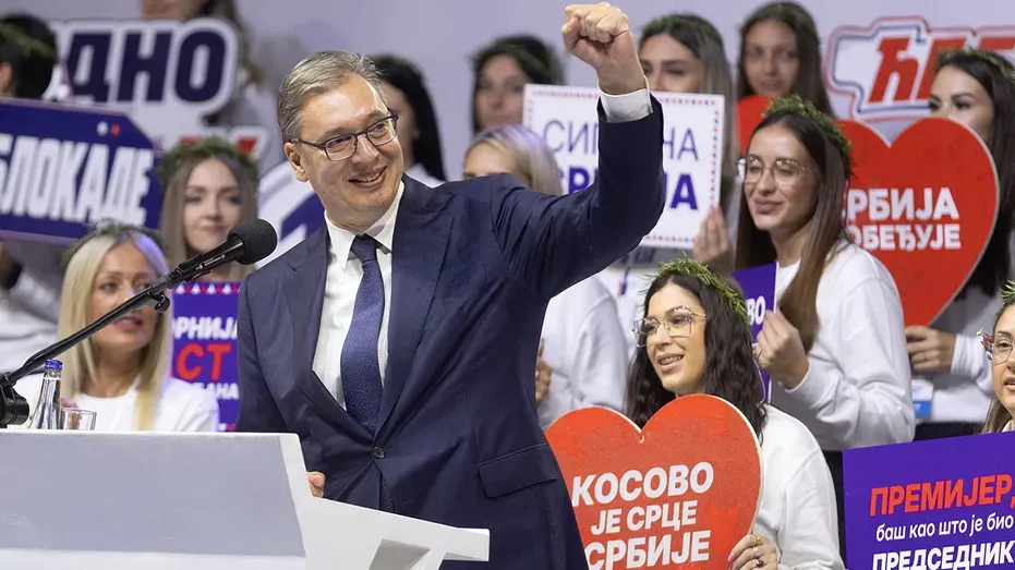 Smiling man in suit raising fist at podium before rally crowd with campaign signs and heart-shaped placards