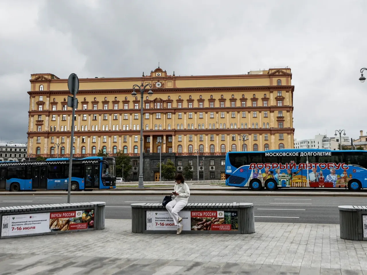 Large yellow Moscow building with clock tower, buses and a woman seated on a bench.