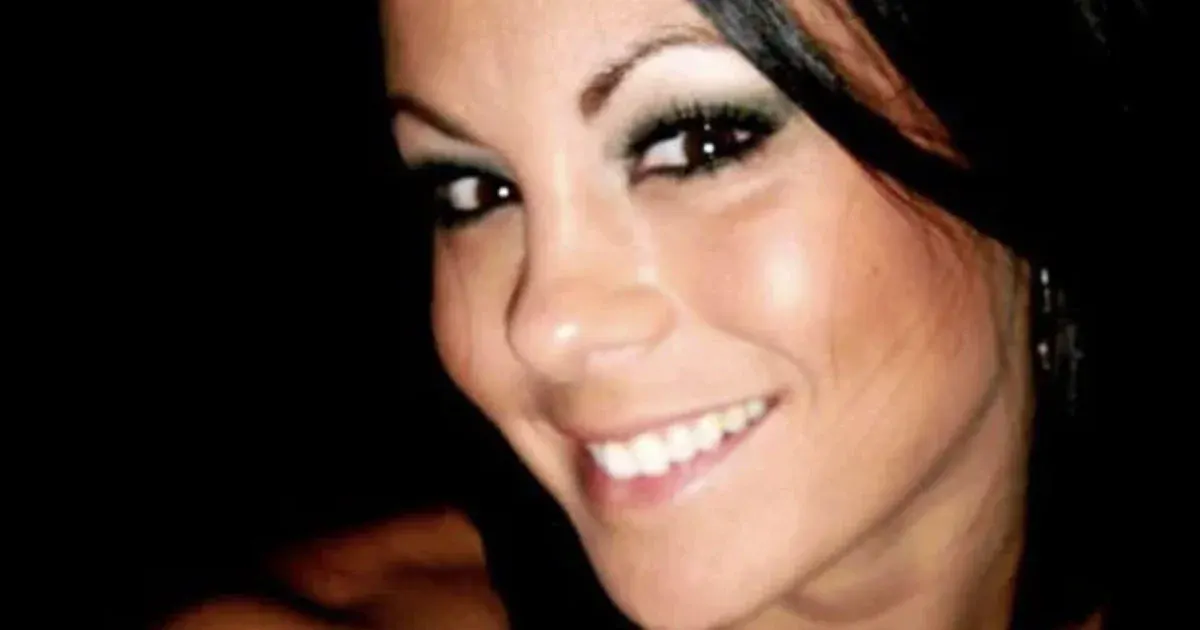 Smiling woman with dark hair and dramatic eye makeup, close-up against a dark background.