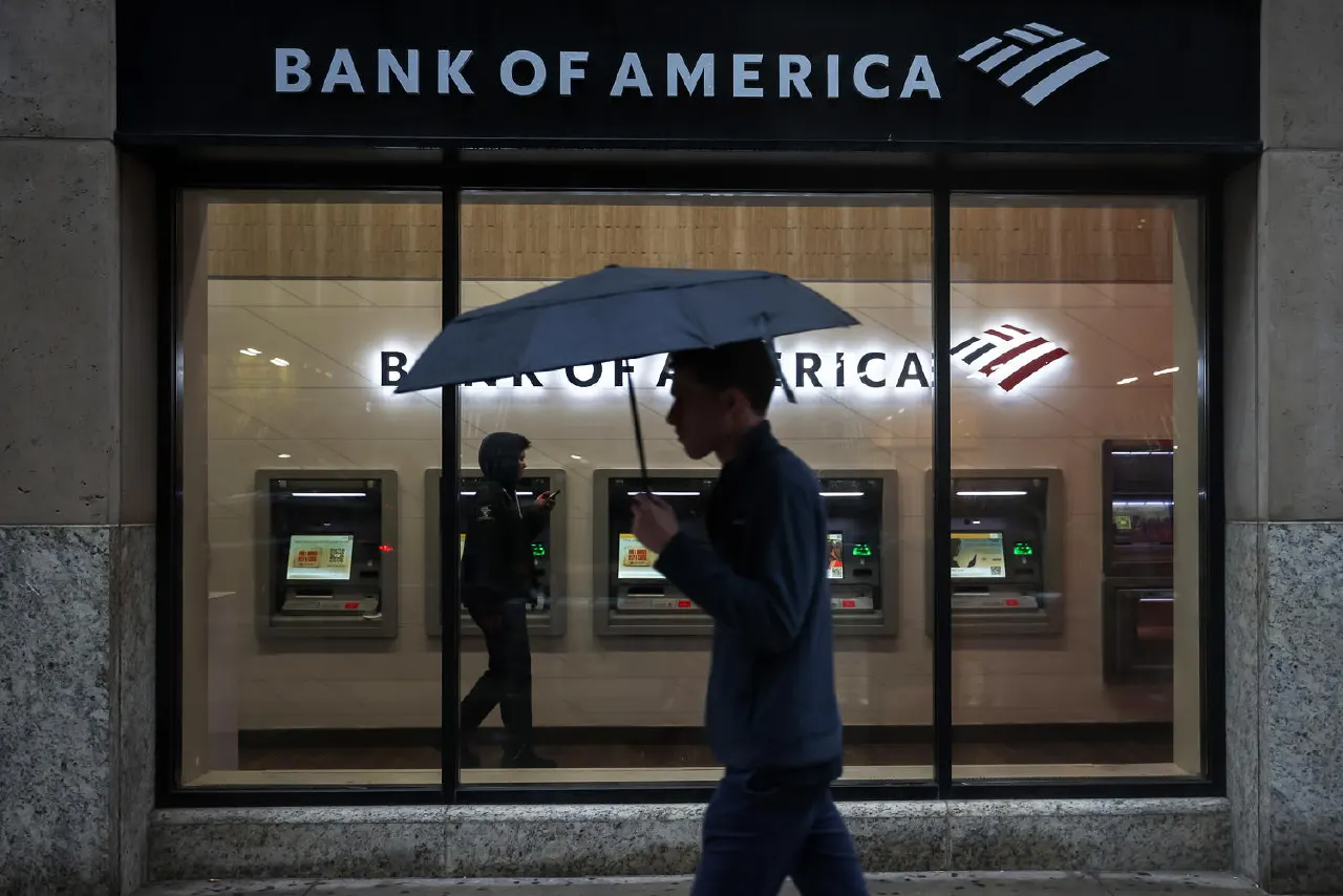 Bank of America branch exterior at dusk with ATMs visible, pedestrians walking past under umbrella.