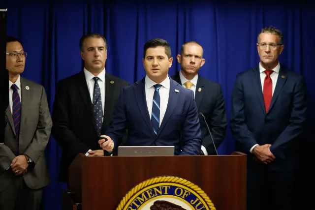 Men in suits at a podium with a Department of Justice seal, speaking at a press event.
