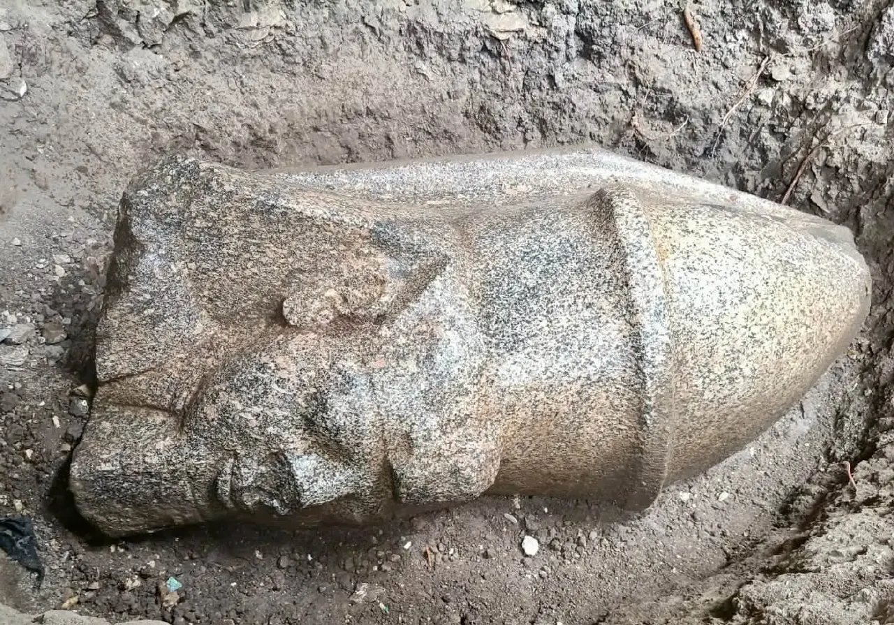Weathered granite statue head partially buried in excavation dirt, face in profile.