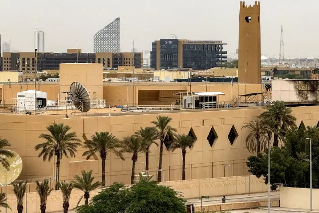 Tan walled compound with palm trees, satellite dish and guard booth, city skyline behind.