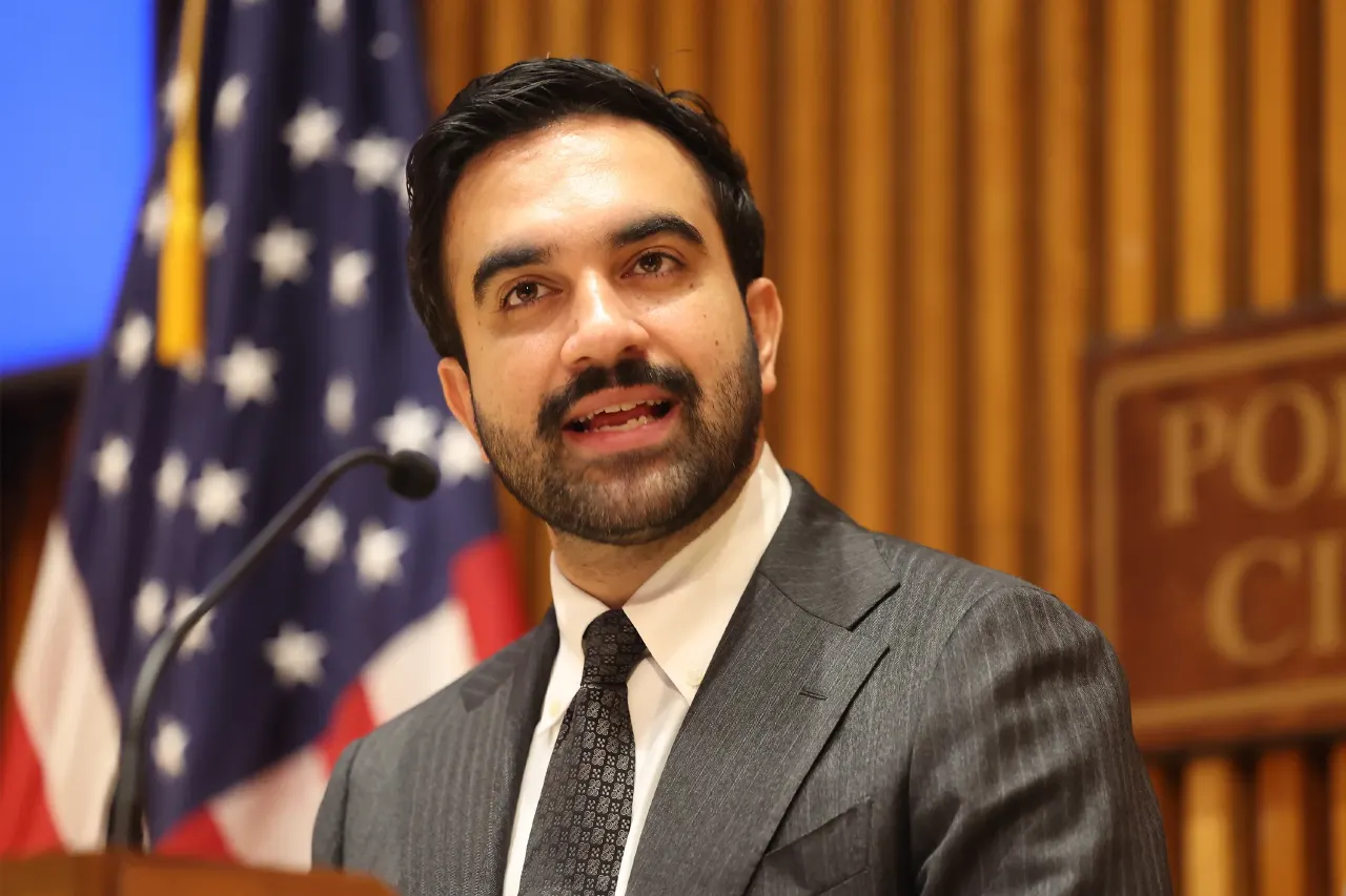 Man speaking at podium with American flag behind him in formal setting.