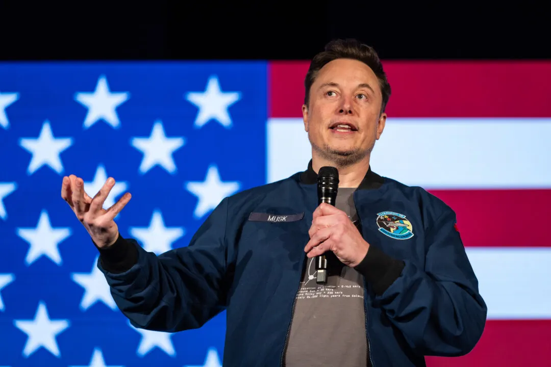 Man in navy jacket speaking into microphone with raised hand, in front of large American flag backdrop.