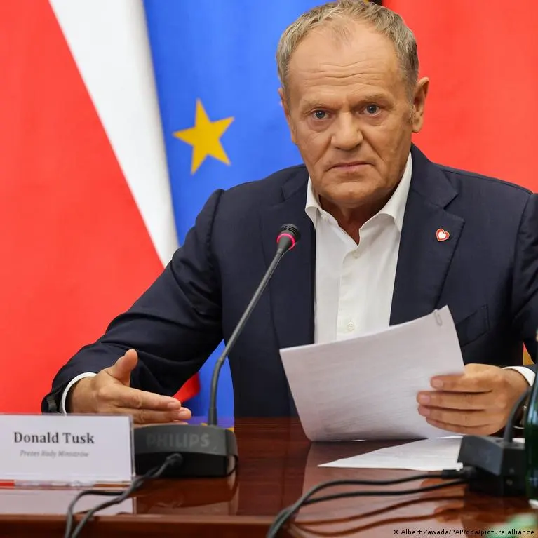 Older man in dark suit seated at microphone, gesturing, holding papers, with flags behind him.