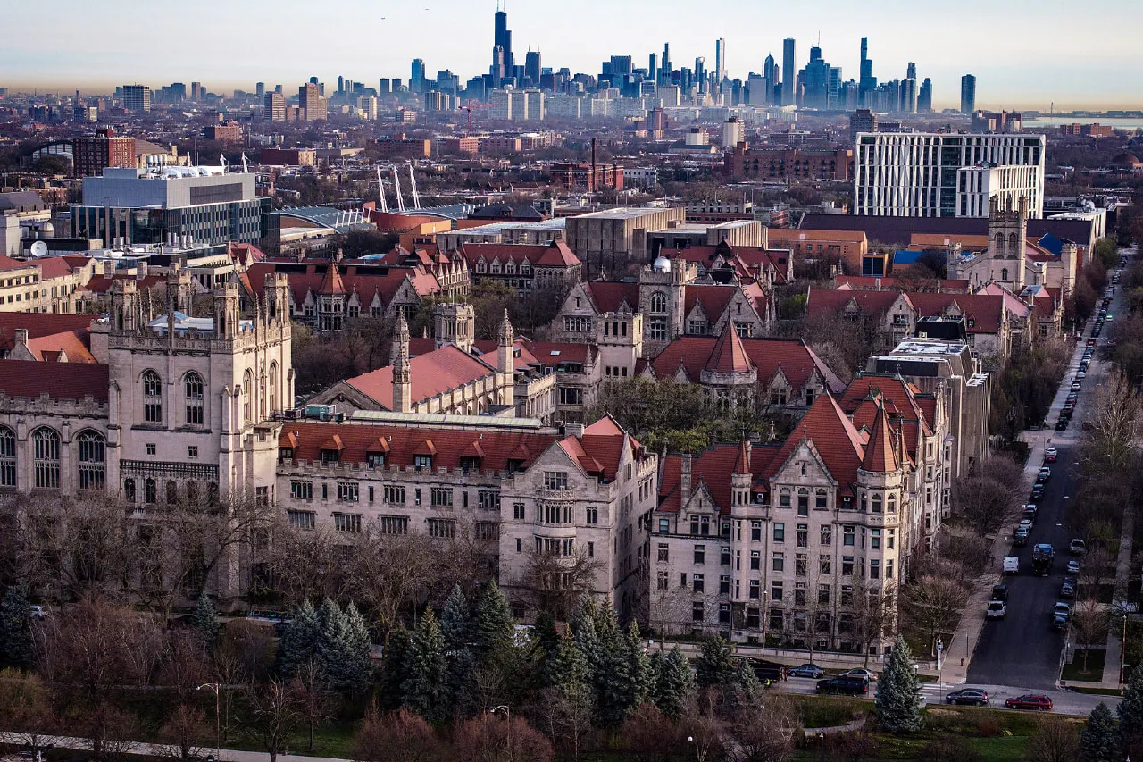 Aerial view of Gothic-style university buildings with red roofs, Chicago skyline behind.