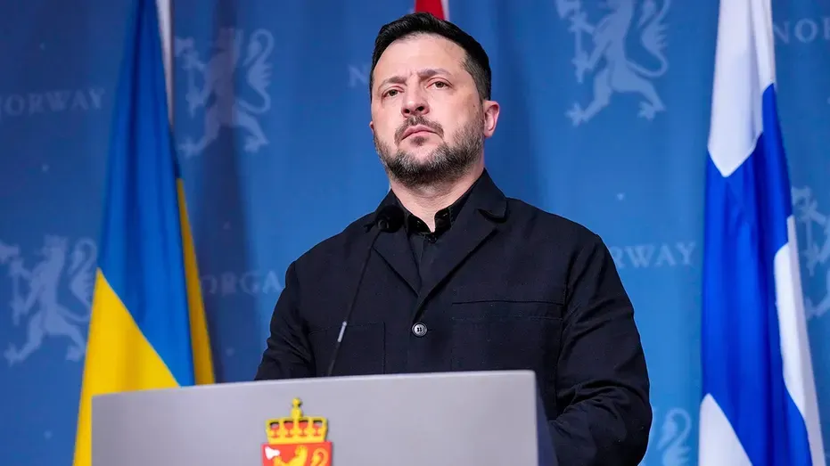 A bearded man in dark jacket speaks at podium before Ukrainian and Norwegian-branded backdrop.