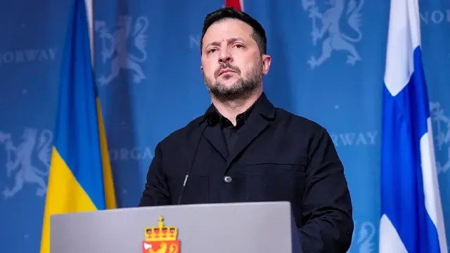 A bearded man in dark jacket speaks at podium before Ukrainian and Norwegian-branded backdrop.