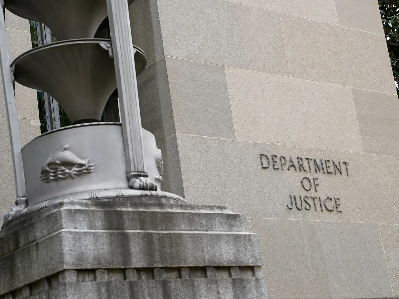 Stone building exterior with 'Department of Justice' lettering beside a carved stone urn pedestal.