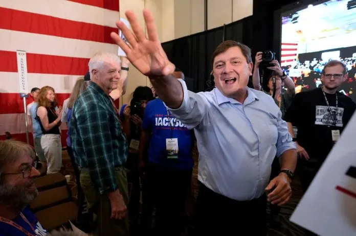 Smiling man waves at a campaign event; a 'Jackson' shirt, American flag and crowd visible.