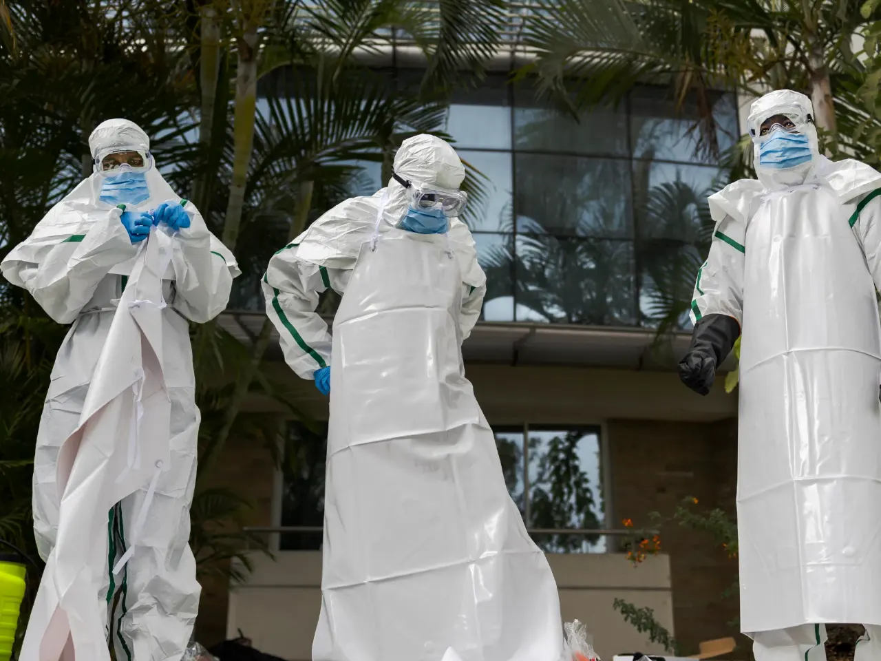Three workers in white protective suits and masks putting on gear outside a building.