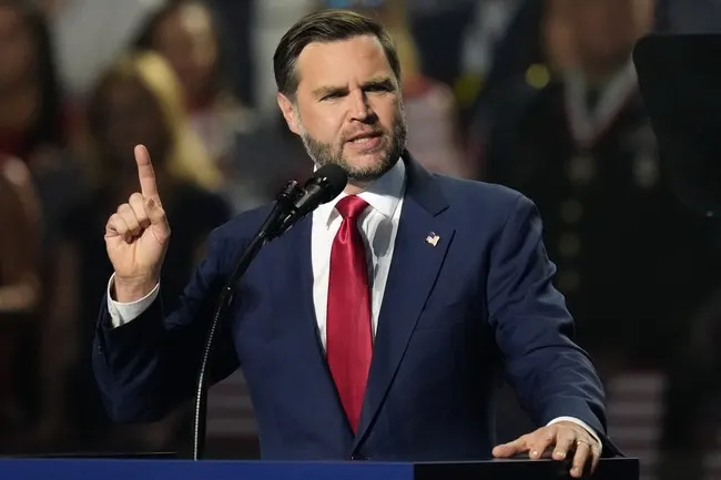 A man in a navy suit and red tie gestures while speaking at a podium with a microphone.