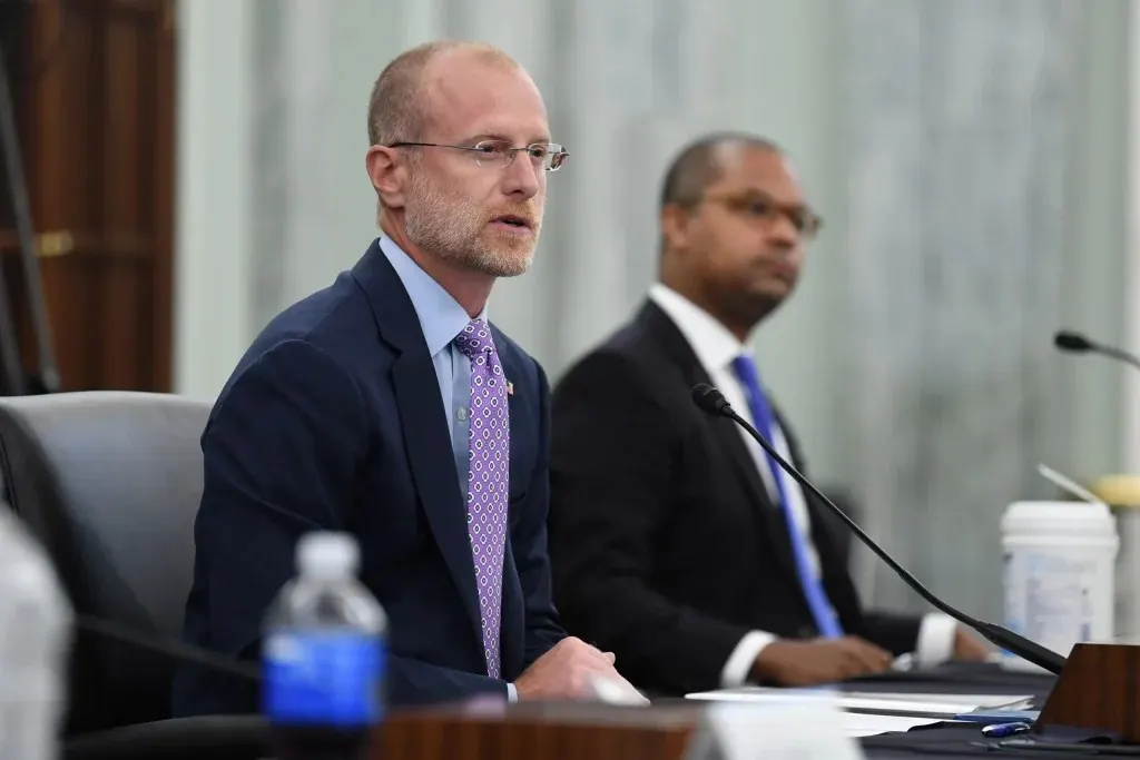 Two men in suits seated at a hearing table with microphones and water bottle.