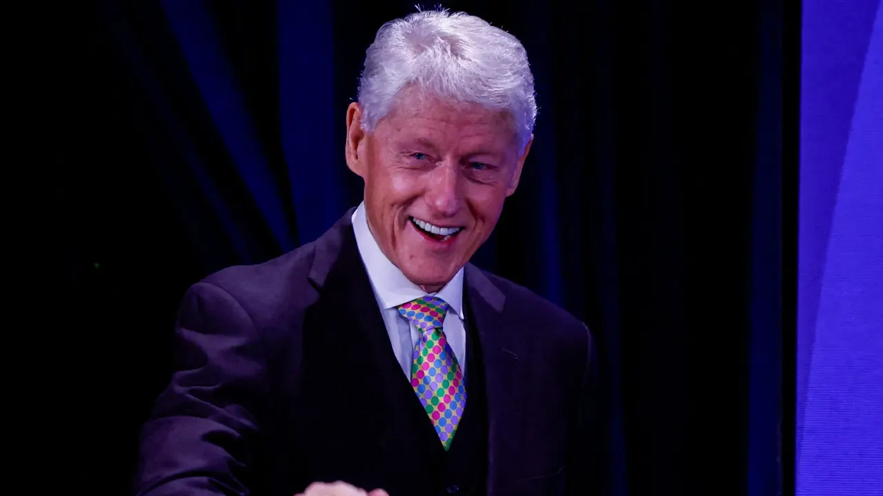 Smiling older man with white hair in dark suit and colorful tie on dark blue stage.