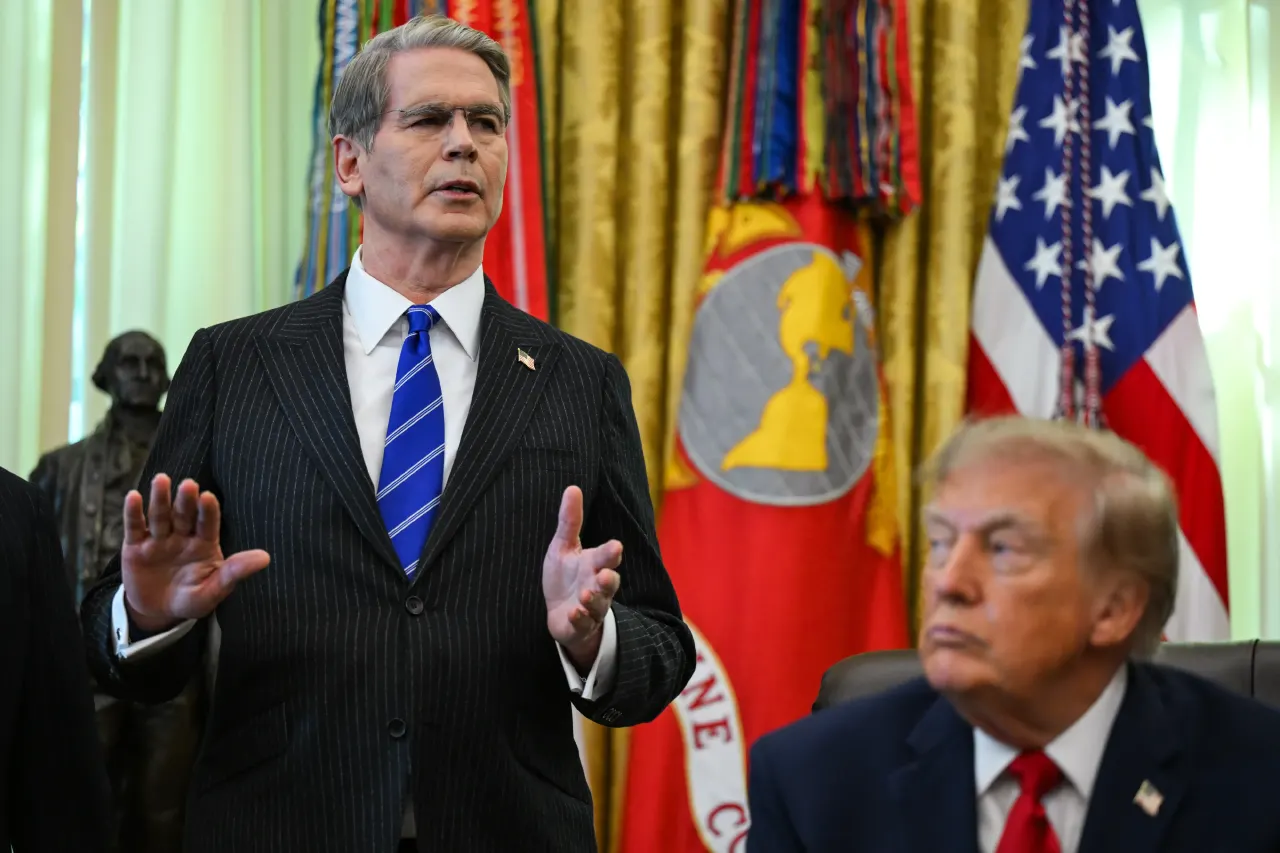 Man in pinstripe suit gestures while speaking, seated man in foreground, American flags and gold curtains behind