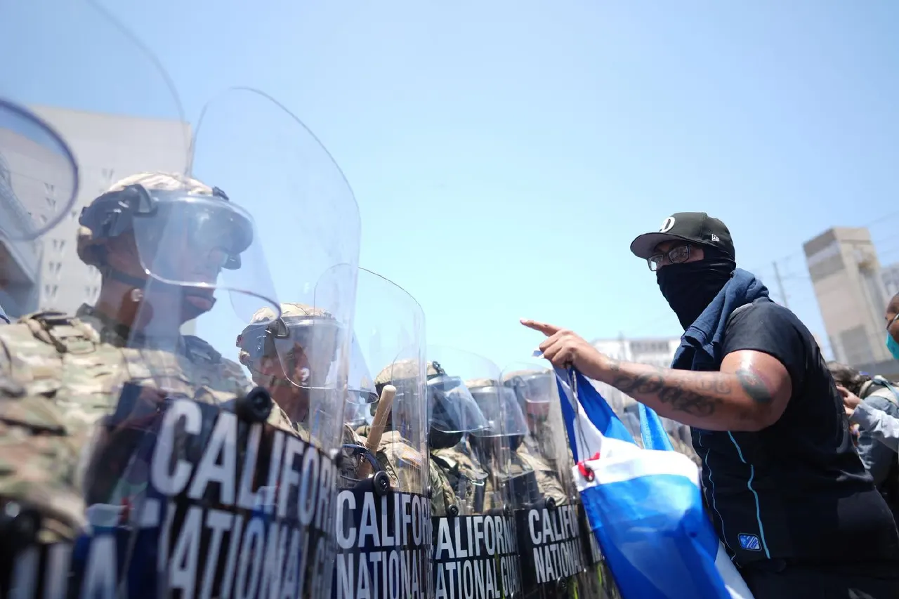 Guard in riot gear with CALIFORNIA NATIONAL GUARD shields faces a masked protester pointing and holding a flag.