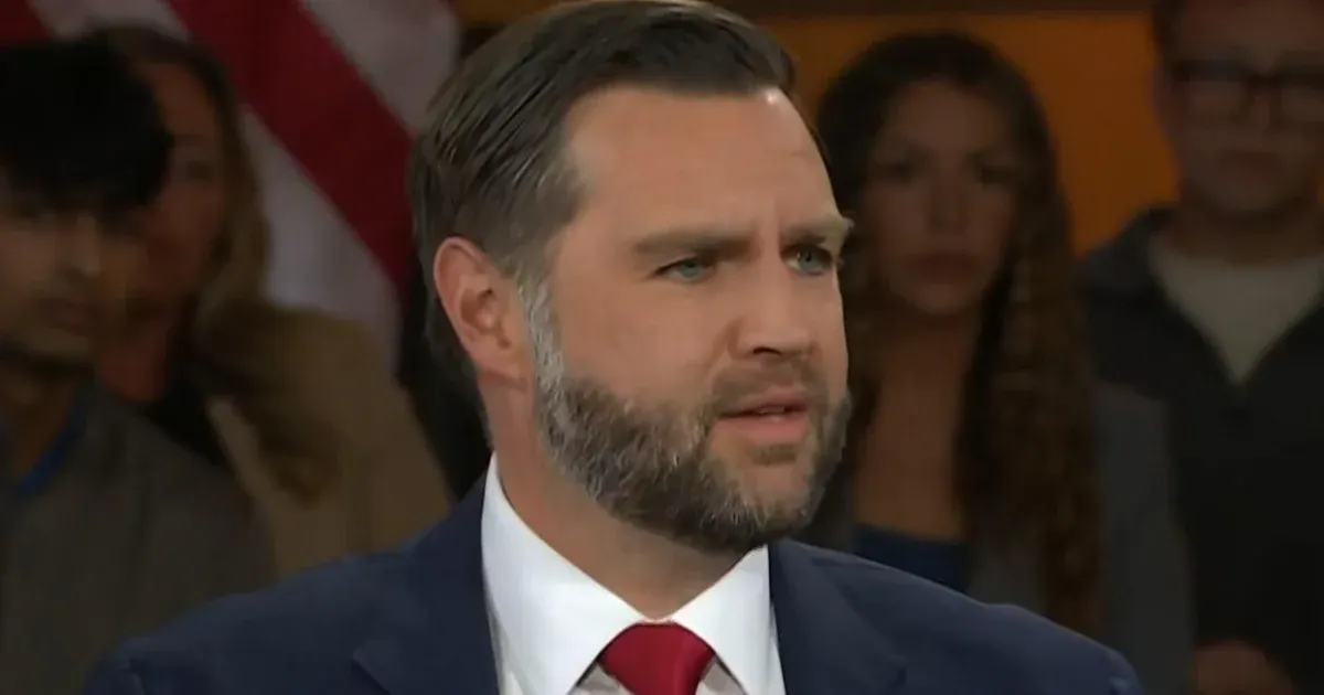 Man in navy suit and red tie speaking at a event, audience and American flag behind him.