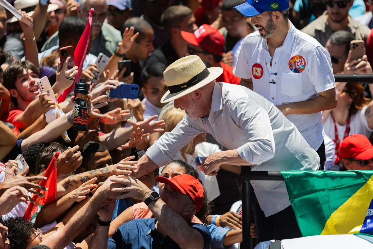 Man in straw hat reaches toward a crowd of supporters holding phones and Brazilian flags at a rally.