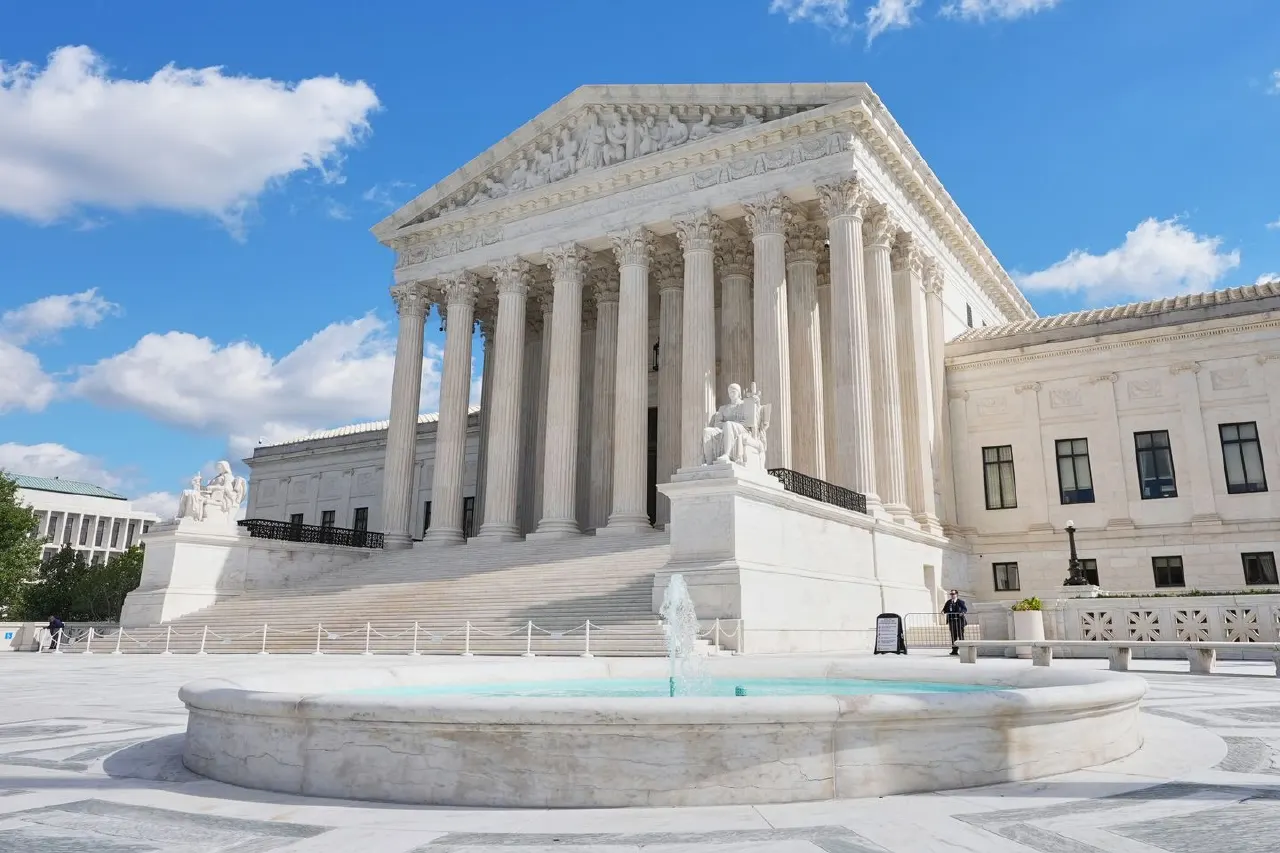 White marble Supreme Court building with columns, fountain, and a distant guard on a sunny day.