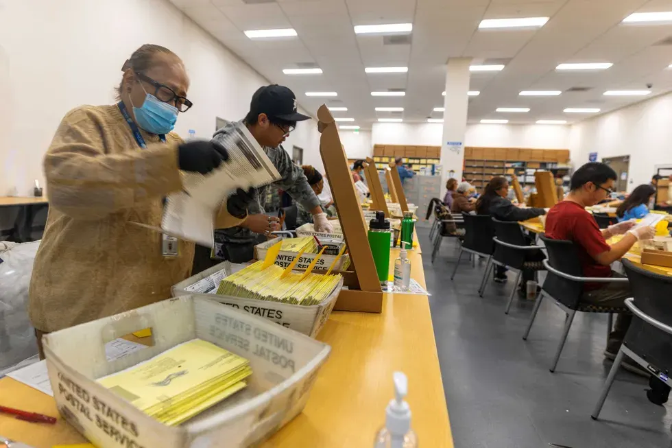 Election workers in a large hall processing yellow ballot envelopes in United States Postal Service trays.