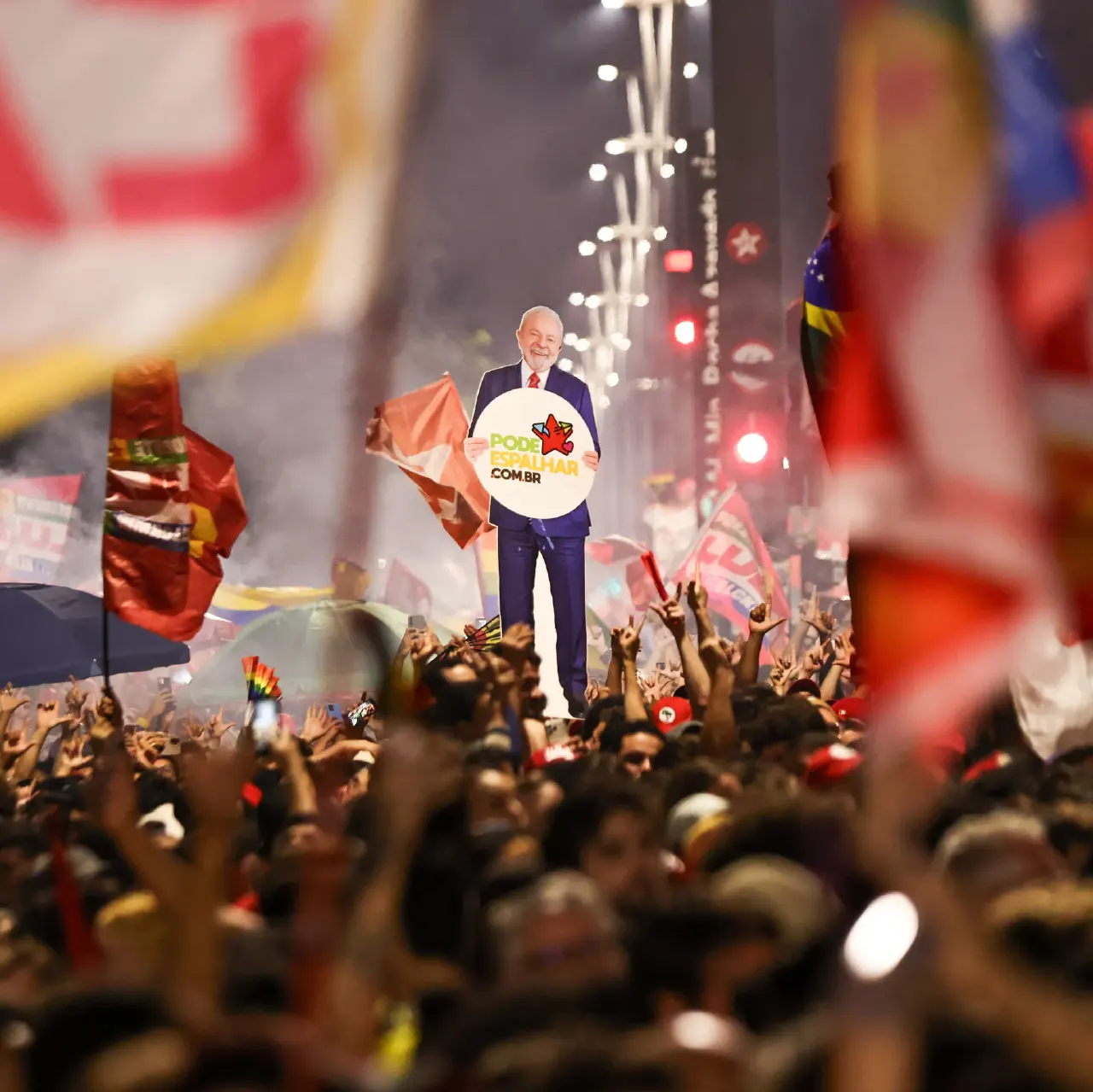 Massive crowd with red flags celebrating; a cutout figure holds a campaign sign reading Pode Cumulhar.