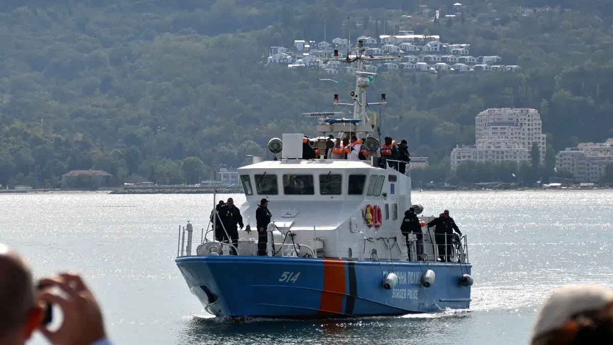 Blue and white coast guard patrol boat with officers on deck, sea and hillside town behind.