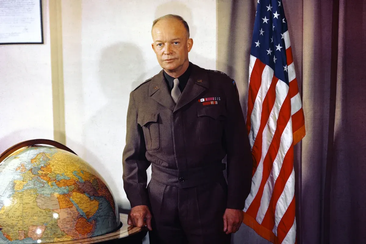 Uniformed man standing beside a vintage globe and a United States flag, in an office setting.