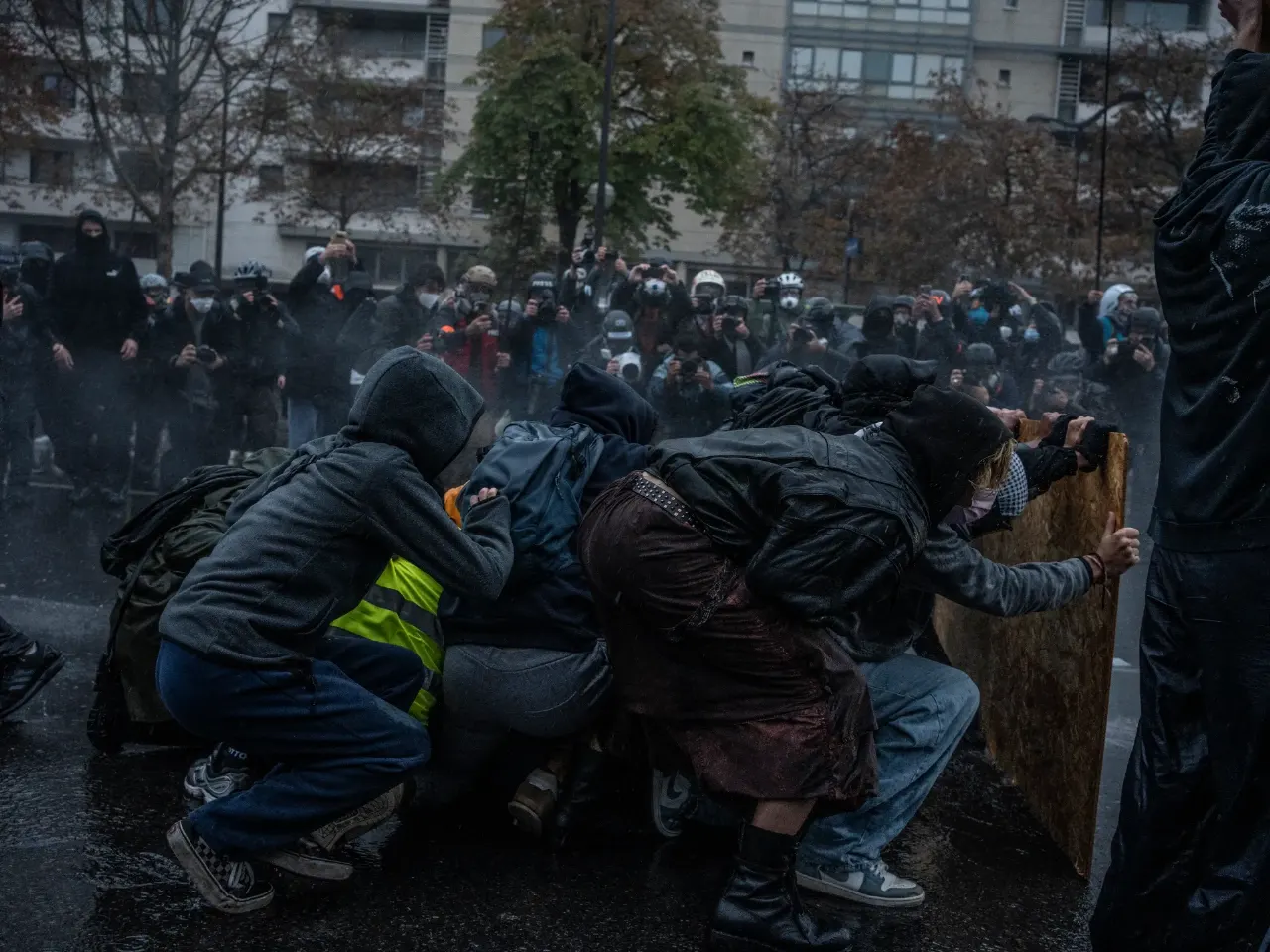 Hooded protesters crouch behind a wooden board as photographers watch amid a street confrontation.
