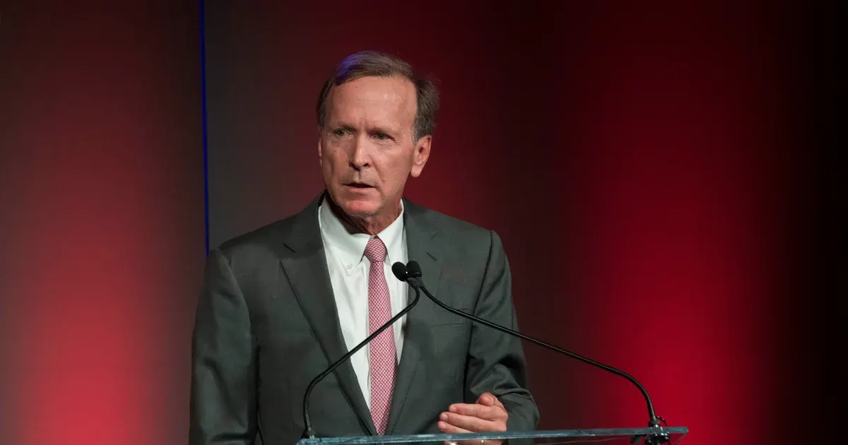 Man in grey suit and pink tie speaking at a glass podium before red backdrop.