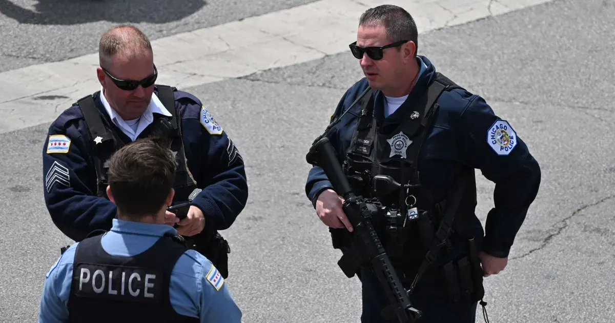 Two uniformed officers in sunglasses and tactical vests, one carrying a rifle, speaking with a third officer.