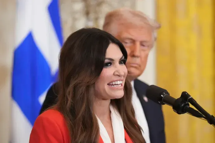 Smiling woman in red jacket at microphone, older man with blurred face behind, blue-and-white flag.