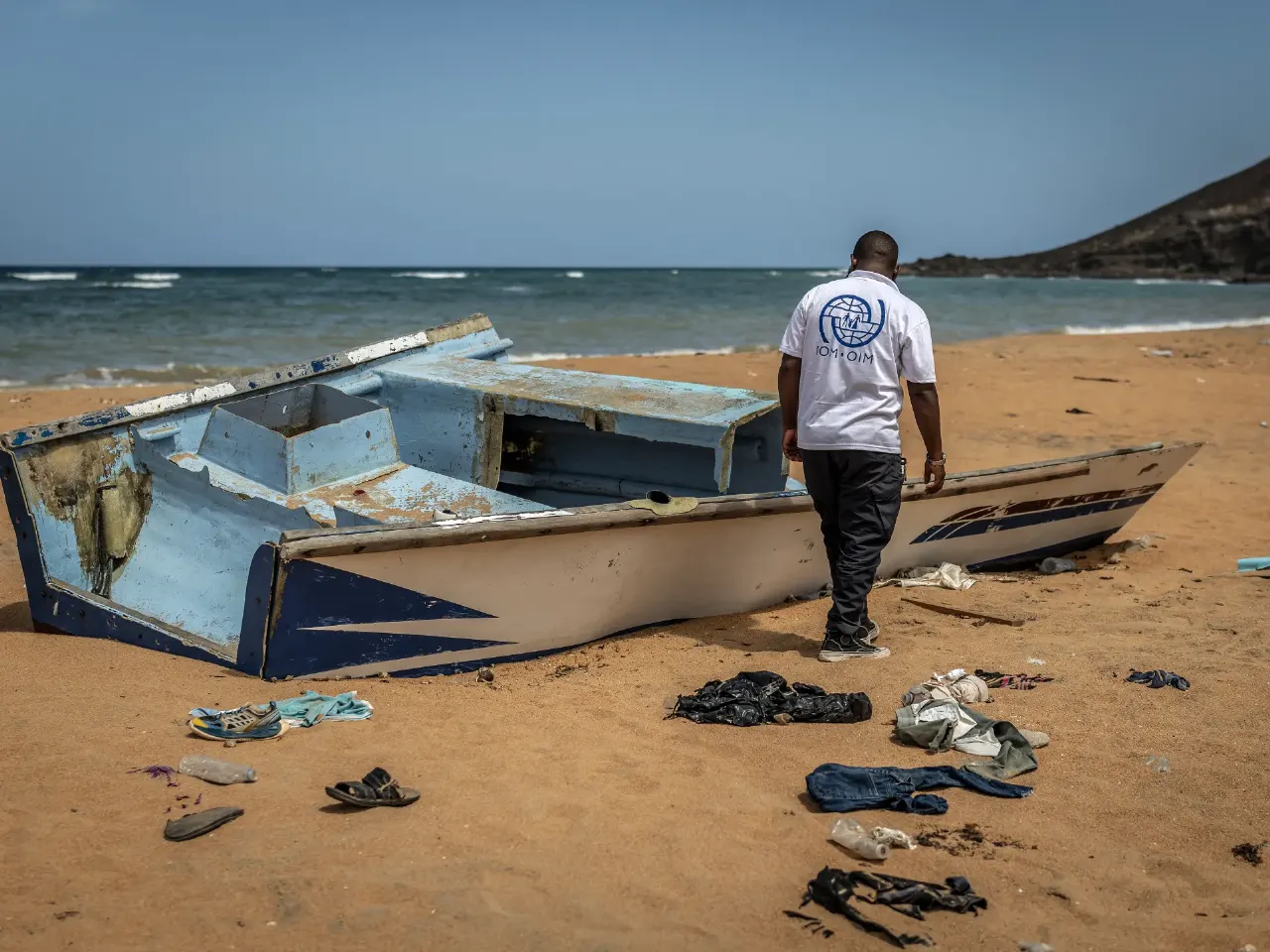 Capsized blue migrant boat on beach with scattered clothing and sandals; man in IOM shirt looks on