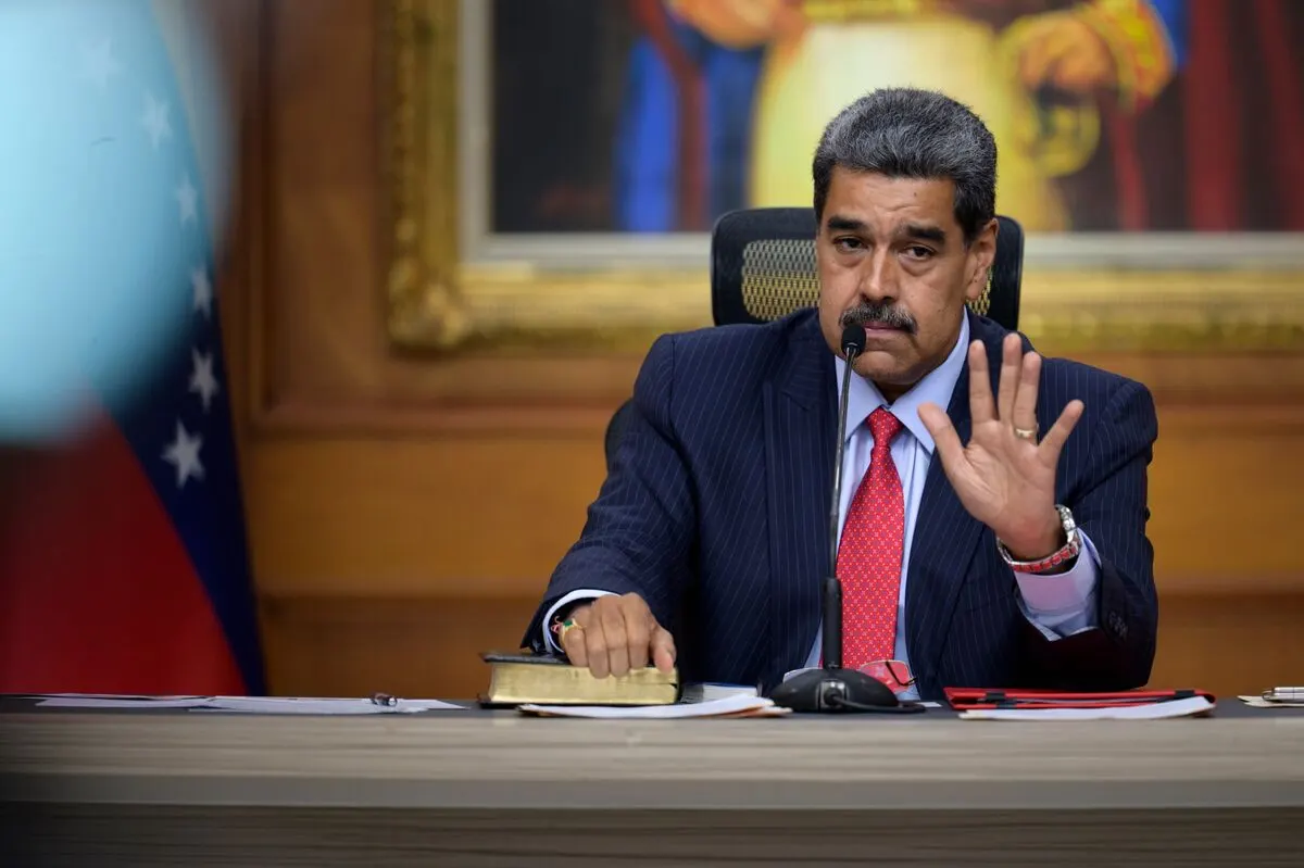 Man in dark suit gestures at a desk before a partially visible national flag, framed portrait behind.