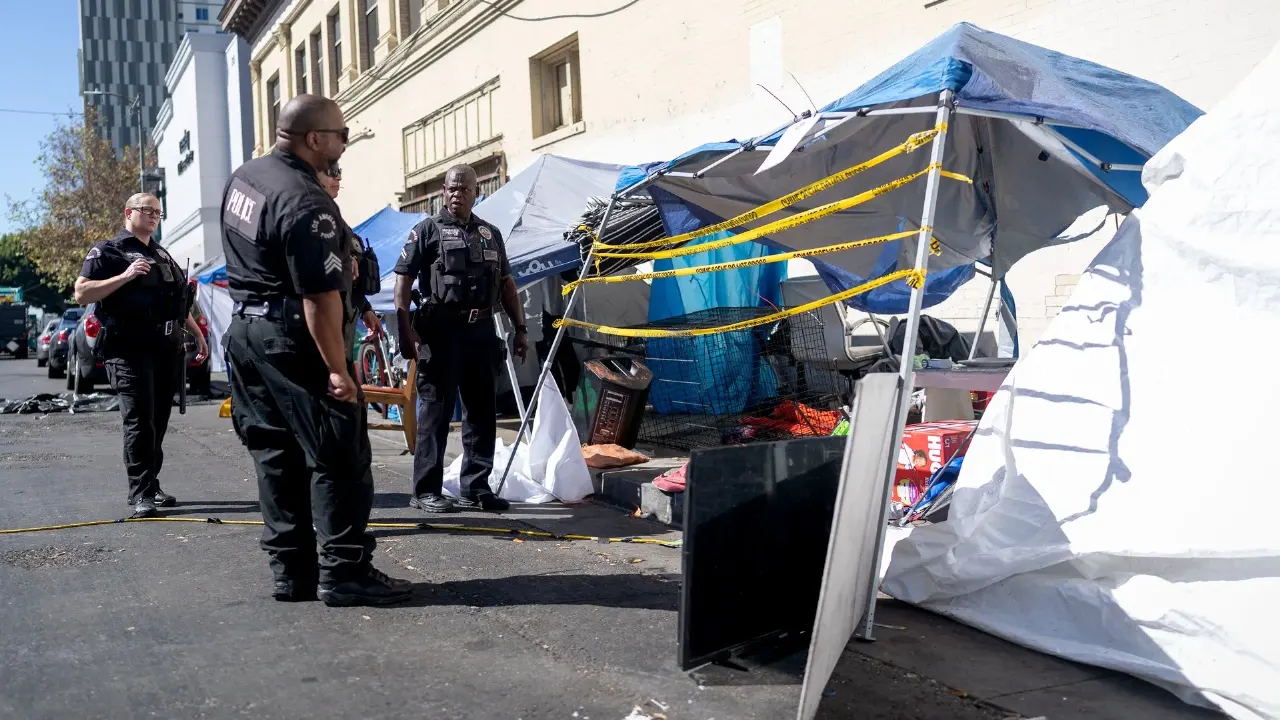 Uniformed officers stand beside a tent encampment cordoned off with yellow tape on a city street.