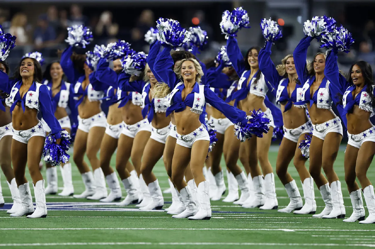 Dancers in blue-and-white star uniforms with pom-poms perform on a football field.