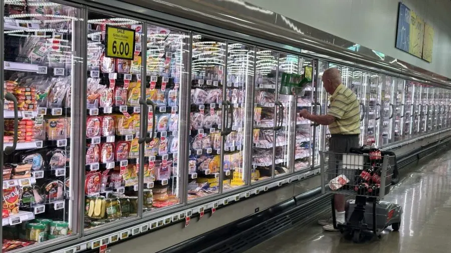 Elderly shopper with cart browsing refrigerated food cases in a supermarket, price sign showing $6.00.