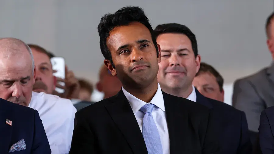Man in dark suit and light tie at an event, surrounded by people, one with a small flag pin.